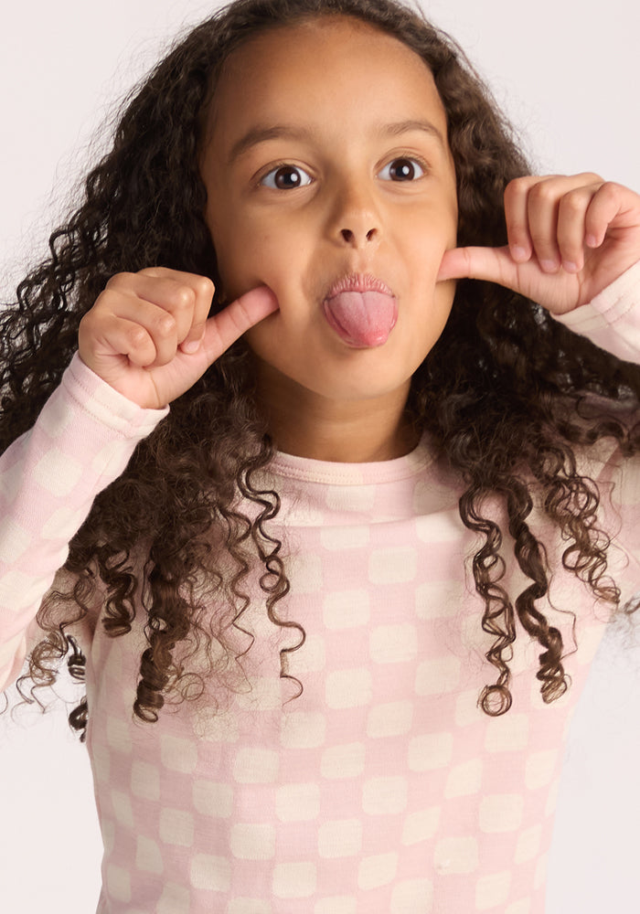 A young girl with long curly hair makes a silly face while wearing the Woolx Kids Explorer Base Layer Top in Hopscotch Pink, standing against a plain background.