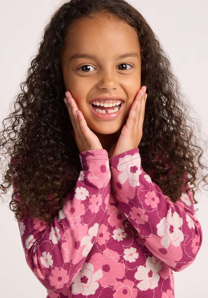 A smiling young girl with curly hair holds her cheeks, wearing the Woolx Kids Explorer Base Layer Top - Rosie Posie, a long-sleeved pink and purple floral Australian Merino wool shirt, against a plain light background.