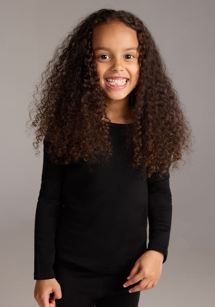 A smiling young child with long, curly brown hair wears the Woolx Kids Explorer Base Layer Top in black and matching black pants, both made from Australian Merino wool, standing against a plain gray background.