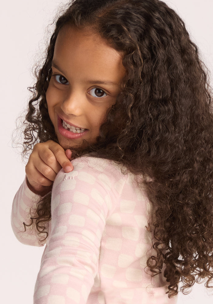 Smiling young girl with long curly brown hair wears the Woolx Kids Explorer Base Layer Top in Hopscotch Pink, pointing to her shoulder while looking back at the camera.