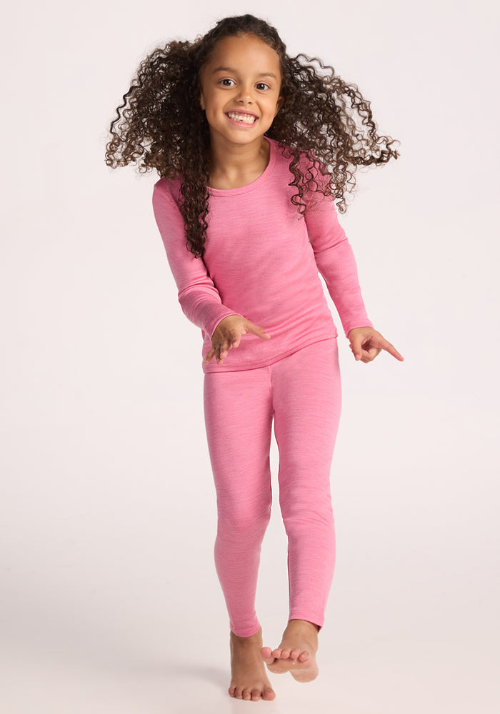 A smiling young girl with long curly hair wears the Woolx Kids Explorer Base Layer Top in Pinky Promise, standing barefoot in motion against a light background. Moisture-wicking Australian Merino wool keeps her comfy all day.