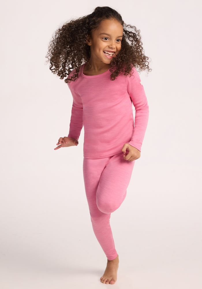 A young girl with curly hair, wearing a long-sleeve pink top and Woolx Kids Explorer Base Layer Bottoms in Pinky Promise, runs barefoot and smiles against a plain light background.