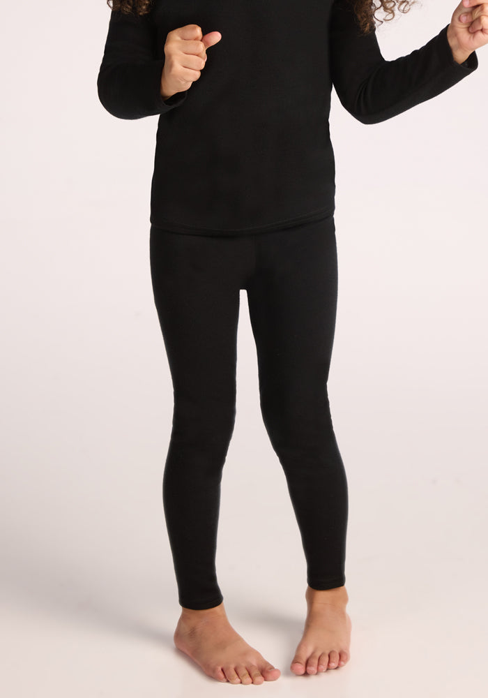 A child stands barefoot on a light background, wearing Woolx Kids Explorer Base Layer Bottoms in black; only the body from shoulders to feet is visible. 