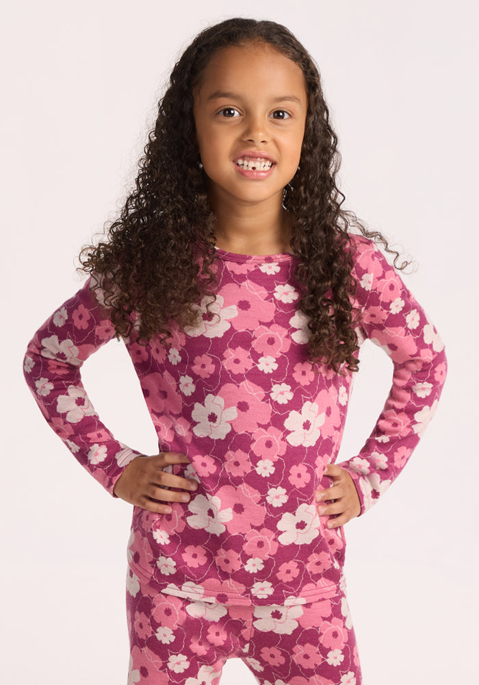 A young girl with long curly hair smiles with her hands on her hips, wearing the Woolx Kids Explorer Base Layer Top in Rosie Posie—a pink and white floral Merino wool top. The background is plain and light-colored.