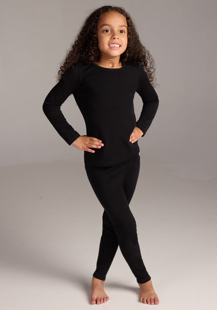 A young child with long curly hair smiles, standing barefoot with hands on hips, wearing the Woolx Kids Explorer Base Layer Top in black against a plain light background.