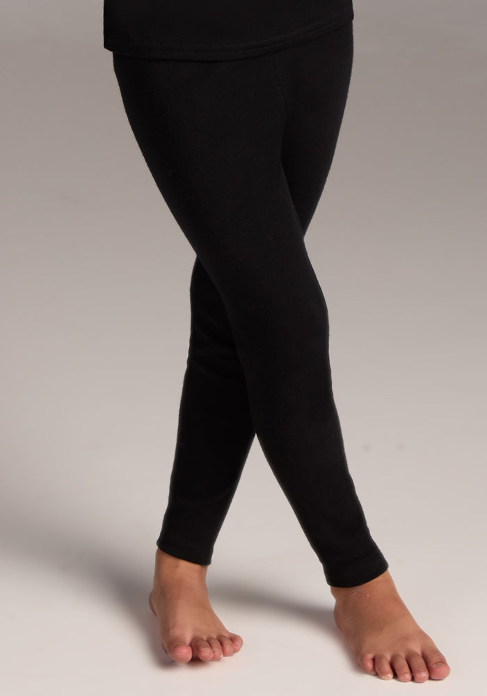 A child models the Woolx Kids Explorer Base Layer Bottoms in black, standing barefoot on a light surface with crossed ankles and outward-pointed toes.