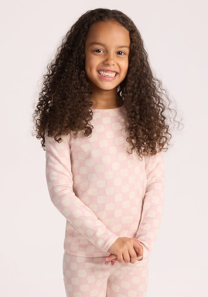 A young girl with long curly hair smiles against a plain background, wearing the Woolx Kids Explorer Base Layer Top in Hopscotch Pink—long-sleeved, cozy, and moisture-wicking—with matching checkered pants for warmth and comfort. 