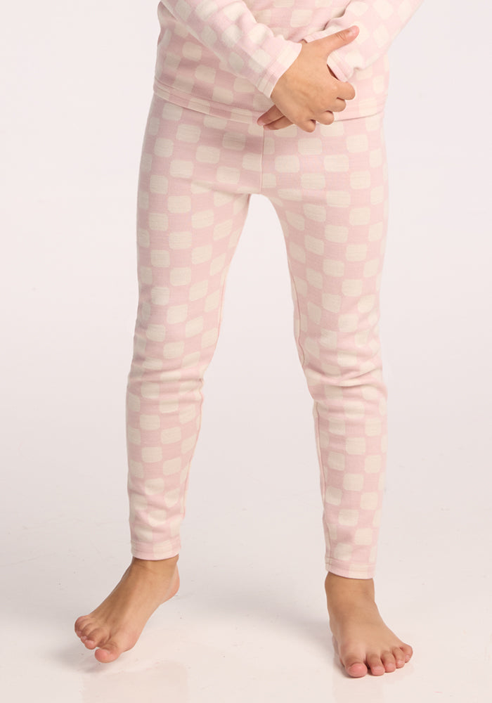 A child models the Woolx Kids Explorer Base Layer Bottoms in Hopscotch Pink, standing barefoot with hands on their lower abdomen. The checkered thermal pants are displayed against a plain white background. 