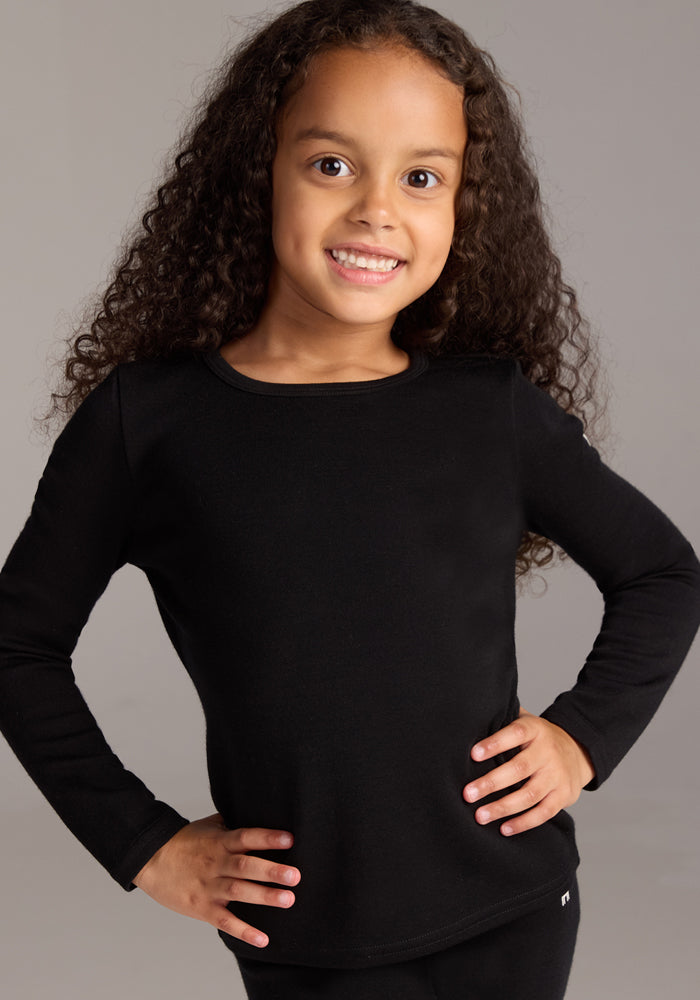 A young girl with long, curly hair smiles confidently, wearing the Woolx Kids Explorer Base Layer Top in black, with her hands on her hips against a neutral background.