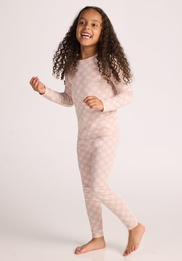 A smiling young girl with long curly hair, barefoot against a plain light background, wears the Woolx Kids Explorer Base Layer Top in Hopscotch Pink—soft, moisture-wicking Australian Merino wool.