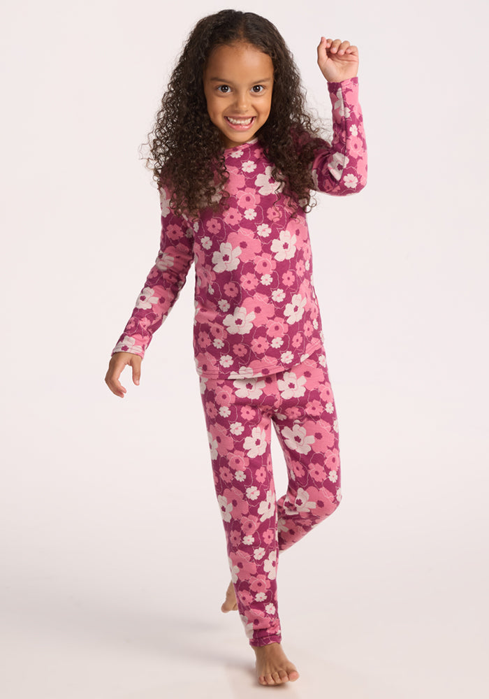 A young girl with curly hair smiles and raises one arm while wearing Woolx Kids Explorer Base Layer Bottoms - Rosie Posie, standing barefoot against a plain light background.