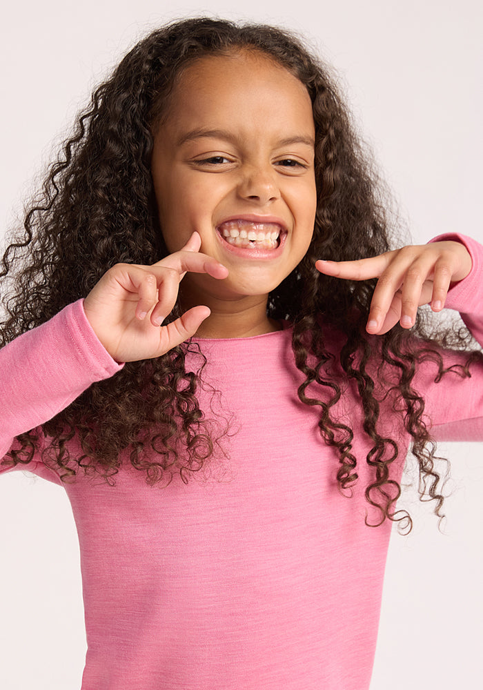 A young child with long curly hair smiles playfully and points at their teeth, wearing the Woolx Kids Explorer Base Layer Top in Pinky Promise, against a plain light background.
