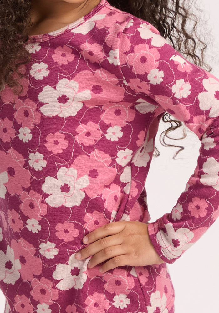 A child stands with one hand on their hip, wearing the Woolx Kids Explorer Base Layer Top in Rosie Posie—a long-sleeve pink and white floral shirt—showing part of their curly hair and face against a white background.