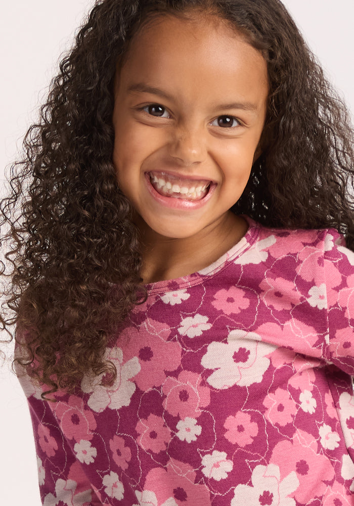 A smiling young girl with long curly hair wears the Woolx Kids Explorer Base Layer Top in Rosie Posie, looking directly at the camera against a light background.