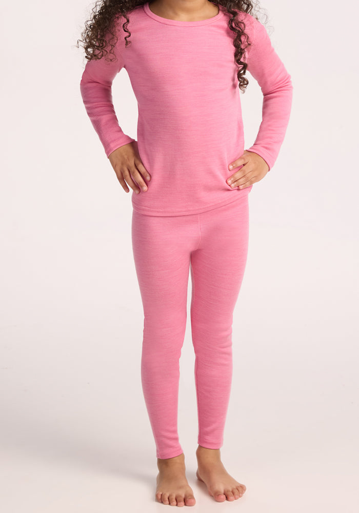 A child stands barefoot wearing Woolx Kids Explorer Base Layer Bottoms - Pinky Promise and a matching long-sleeve pink top, hands on hips, face not visible.