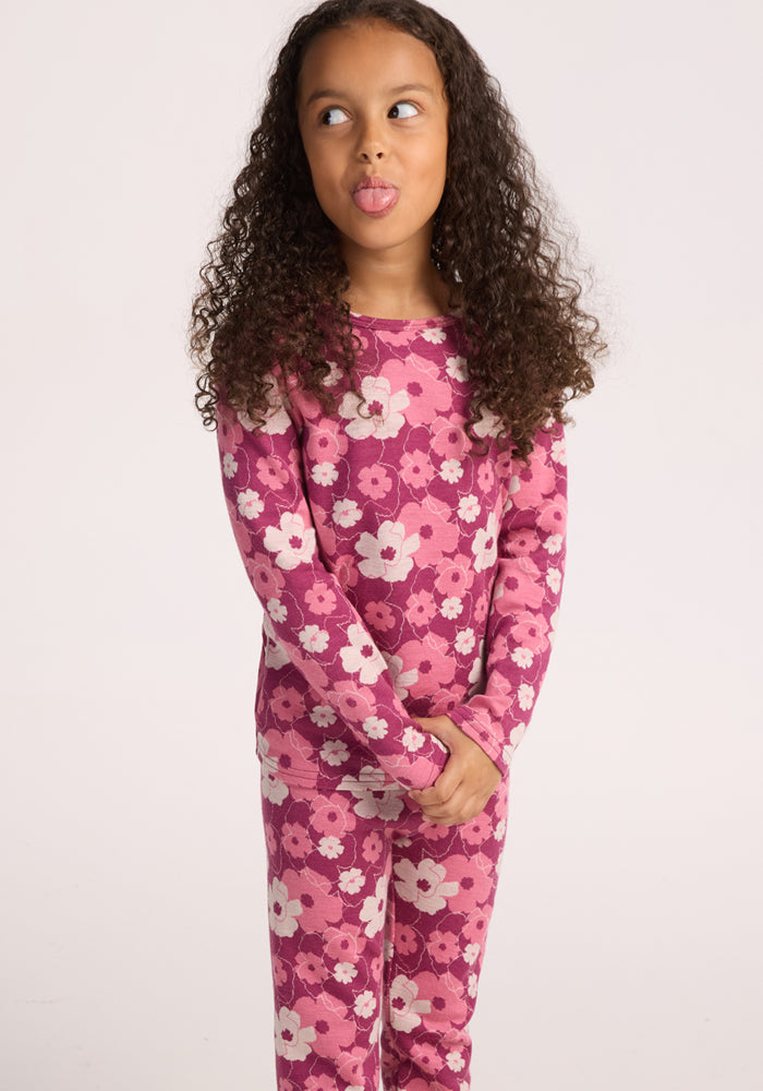 A young girl with curly hair smiles playfully and sticks out her tongue while wearing Woolx Kids Explorer Base Layer Bottoms - Rosie Posie, standing against a light background with her hands clasped in front of her.