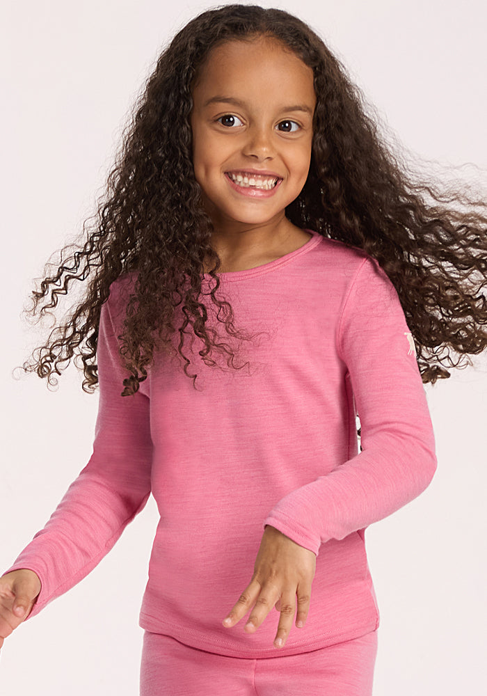 A young child with long curly hair smiles widely while wearing the Woolx Kids Explorer Base Layer Top in Pinky Promise, standing against a plain light background.