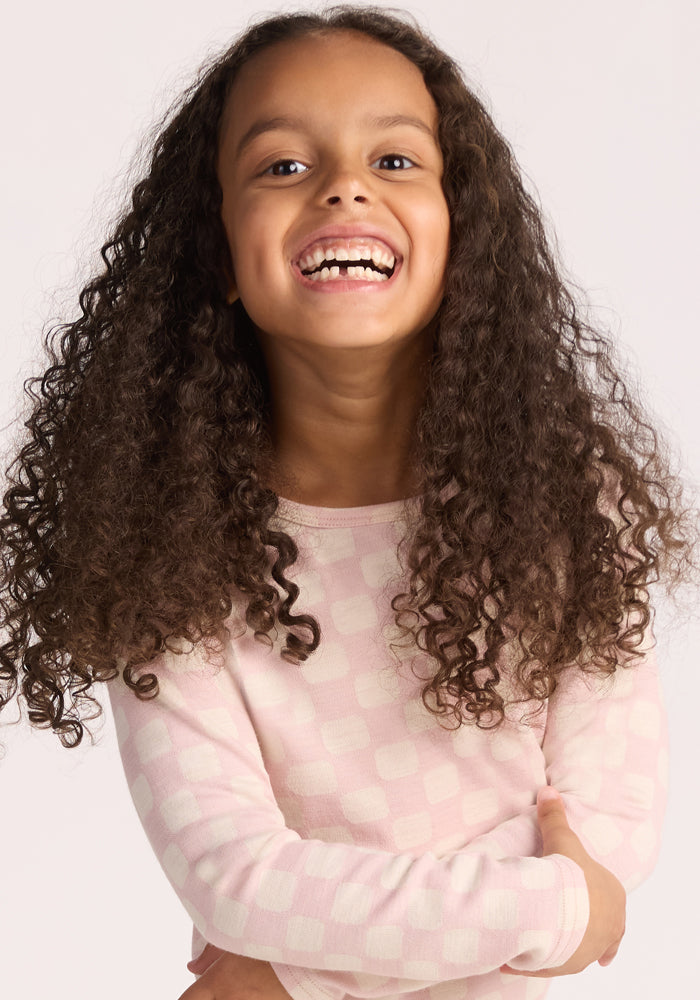 A young girl with long, curly brown hair smiles brightly, crossing her arms. She wears the Woolx Kids Explorer Base Layer Top in Hopscotch Pink, a light pink merino wool top with a subtle checkered pattern. The background is plain and light-colored.