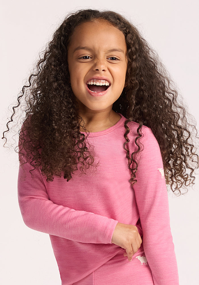 A young girl with long, curly brown hair laughs while wearing the Woolx Kids Explorer Base Layer Top in Pinky Promise, standing against a plain light background.