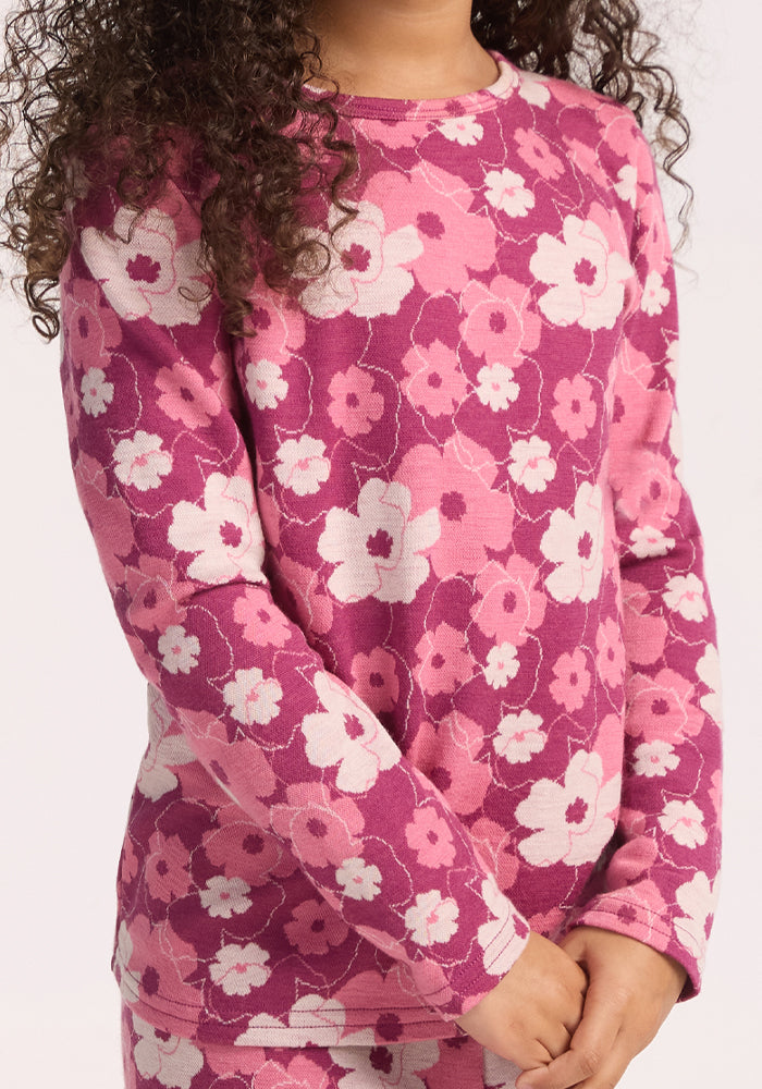 A child with curly hair wears the Woolx Kids Explorer Base Layer Top - Rosie Posie, a warm, long-sleeve pink and magenta Australian Merino wool shirt with a white and light pink floral pattern, standing with arms relaxed.