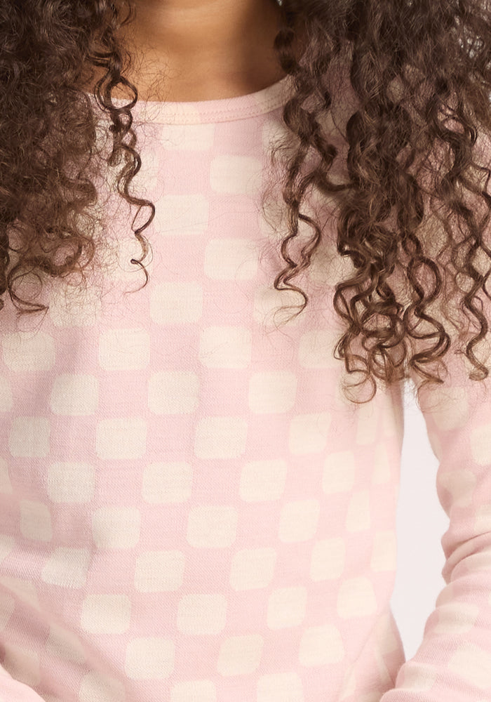 A close-up of someone with curly hair wearing the Woolx Kids Explorer Base Layer Top in Hopscotch Pink; its moisture-wicking fabric features a subtle white checkered pattern, and the person's face is mostly out of frame.