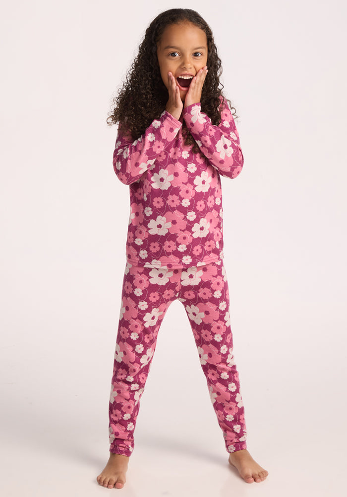 A young girl with curly hair stands barefoot, smiling with her hands on her cheeks. She wears Woolx Kids Explorer Base Layer Bottoms in the Rosie Posie print against a plain light background.