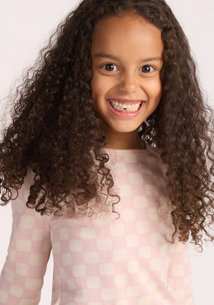 A smiling child with long, curly hair wears the Woolx Kids Explorer Base Layer Top in Hopscotch Pink, standing against a plain light background—ideal for all-day comfort with moisture-wicking merino wool.