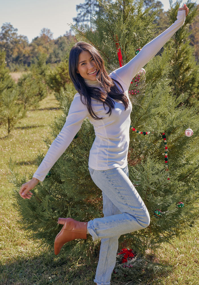 A woman wearing the Woolx Peyton Turtleneck in Buttercream and jeans smiles at the camera, striking a playful pose with arms outstretched by a decorated outdoor pine tree.
