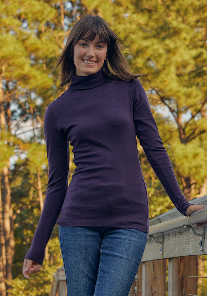 A woman with straight brown hair, wearing the Woolx Peyton Turtleneck in Deep Plum and blue jeans, smiles on a wooden bridge outdoors surrounded by green trees.