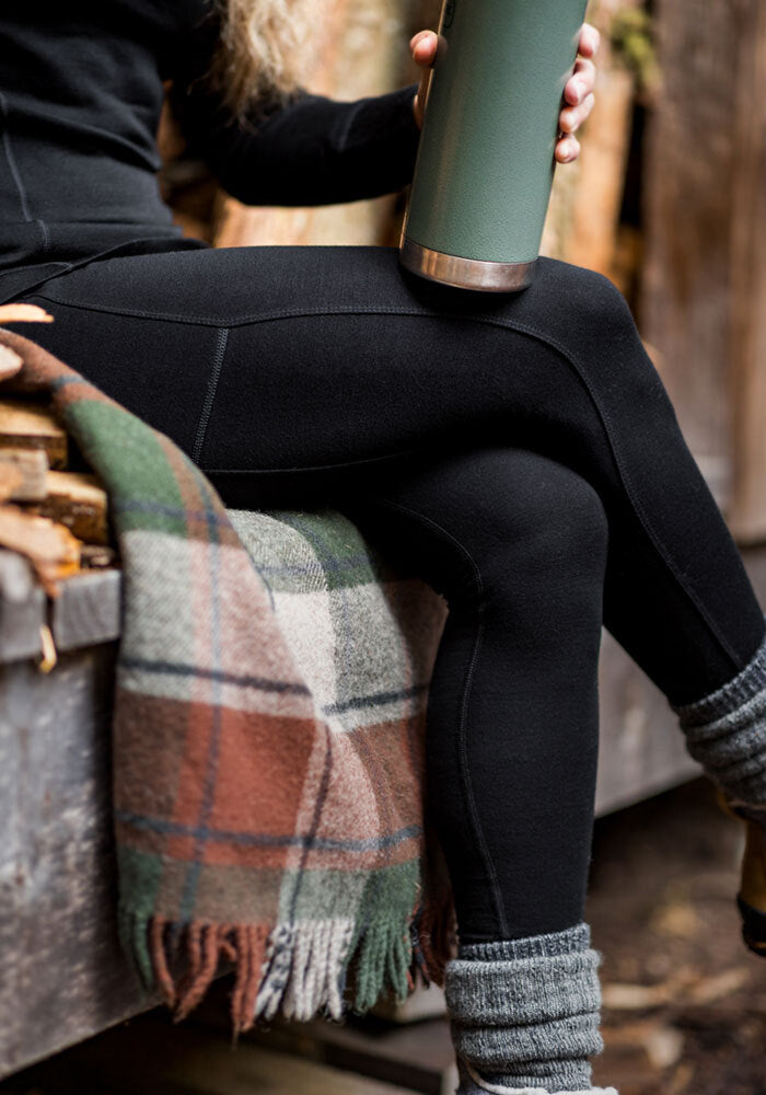 Person in Woolx Piper Pocket Tall - Black leggings sits on a bench with a plaid blanket, holding a green thermos.