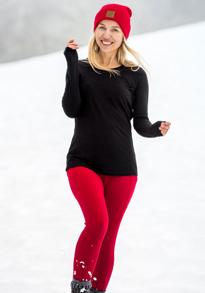 Smiling woman in Woolx Piper Pocket Leggings - Maraschino Cherry, black top, red hat, walking on snowy ground.