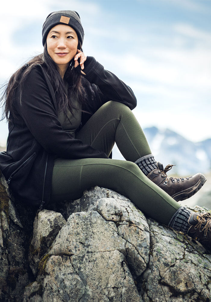 Woman sits on a rock smiling, with Woolx Piper Pocket Tall Duffel Bag; mountains and clouds in the background.