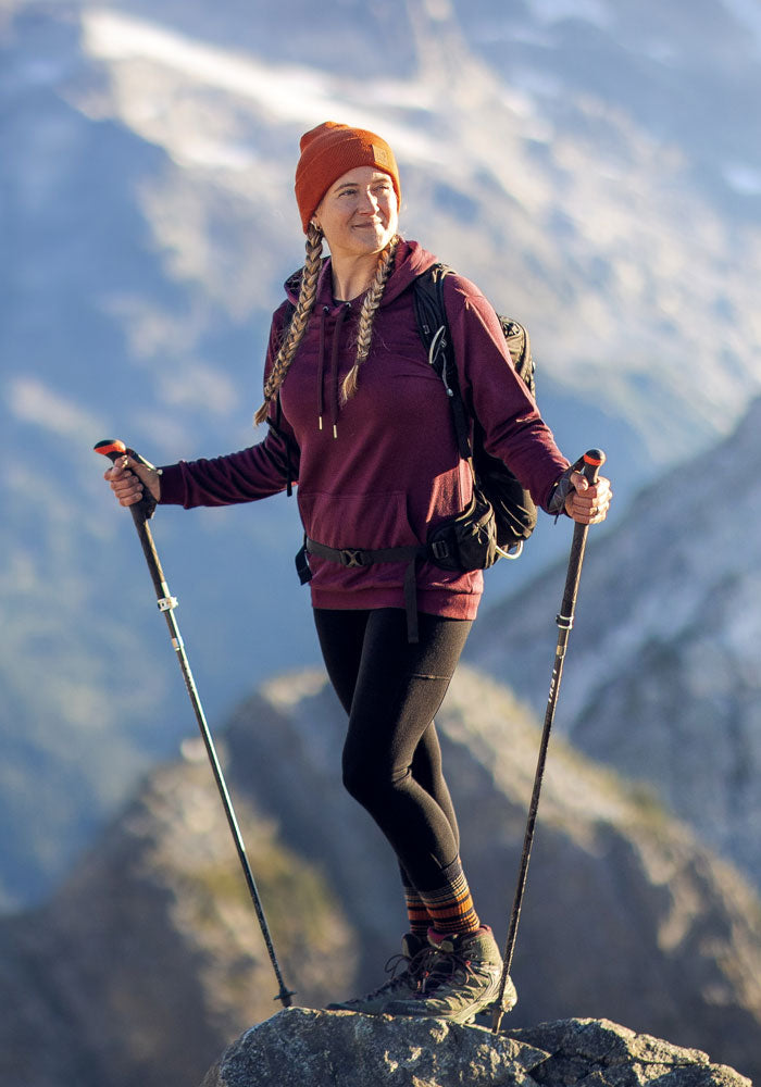 A woman in hiking gear stands on a rocky peak, holding trekking poles. She wears Woolx Piper Pocket Leggings in black, a maroon hoodie, orange beanie, and backpack. Snow-capped mountains and a blurred valley form the backdrop.