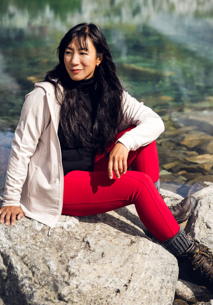 Woman in Woolx Piper Pocket Leggings - Maraschino Cherry sits by clear water, looking off to the side outdoors.