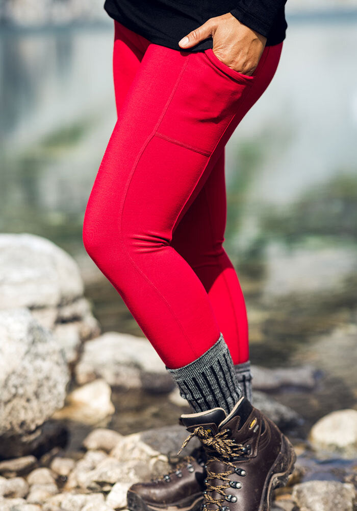 Person in Woolx Piper Pocket Leggings - Maraschino Cherry, gray socks, and boots stands on rocks near water.