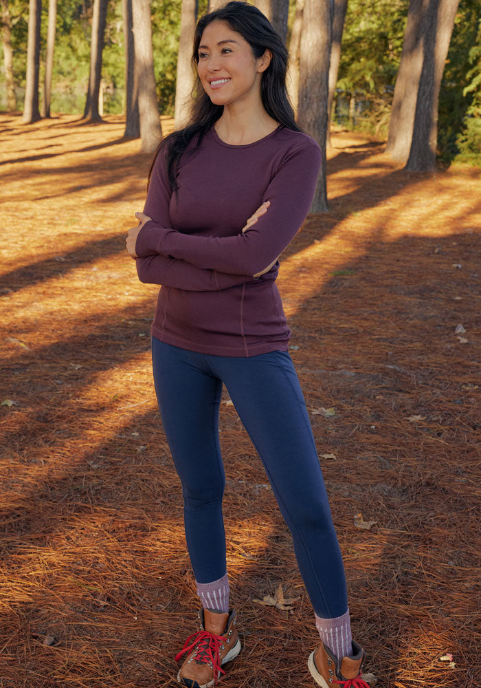 A woman stands on a pine needle-covered forest floor, smiling with arms crossed. She wears Woolx Piper Pocket Leggings in Deep Navy and a maroon long-sleeve top as sunlight filters through tall trees behind her.