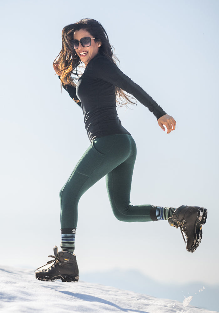 A woman in Woolx Piper Pocket Petite - Deep Timber leggings joyfully jumps on snow, wearing sunglasses, a black long-sleeve top, and hiking boots under a clear sky.