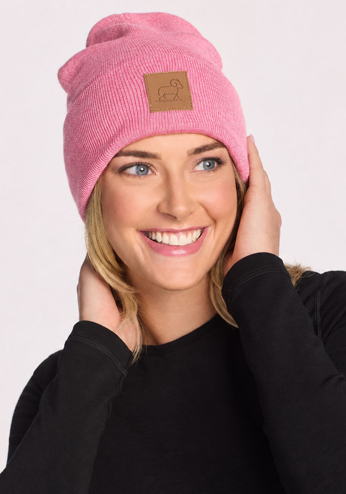 Smiling woman in Woolx Baylor Beanie - Pinky Promise and black shirt, holding the hat’s sides against a light background.