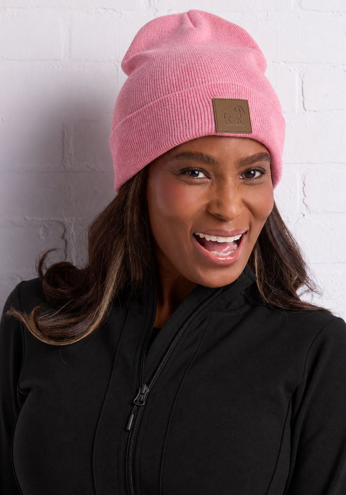Woman in a Woolx Baylor Beanie - Pinky Promise and black jacket smiles before a white brick wall.
