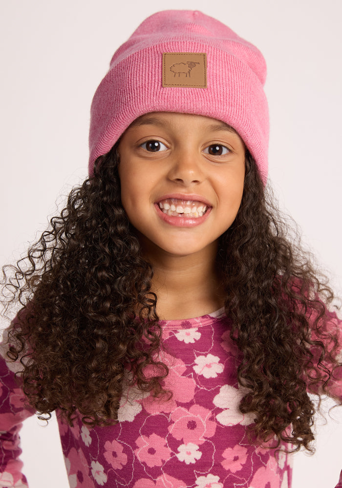 A young girl with long curly hair smiles at the camera, wearing the Woolx Kids Baylor Beanie in Pinky Promise against a plain light background.