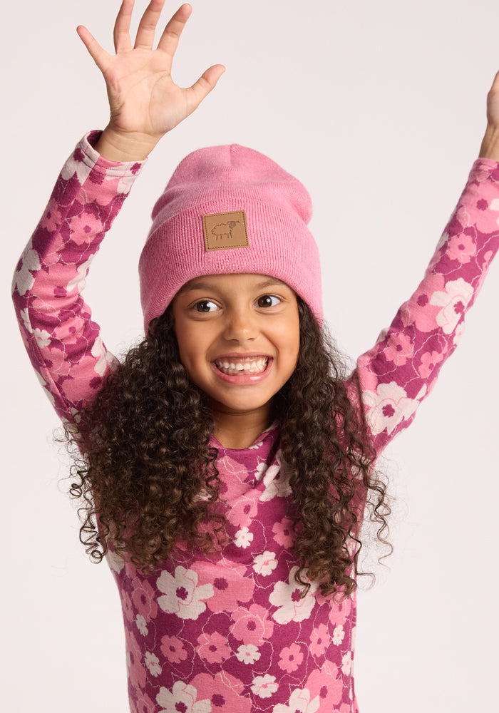 A young girl with long curly hair smiles brightly, wearing the Woolx Kids Baylor Beanie in Pinky Promise and a pink floral shirt, raising both arms against a plain light background.