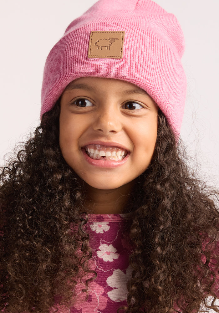 A young girl with long curly hair wears the Woolx Kids Baylor Beanie in Pinky Promise—a pink knit hat with a sheep patch—and a pink floral shirt, smiling and looking slightly to the side against a light background.