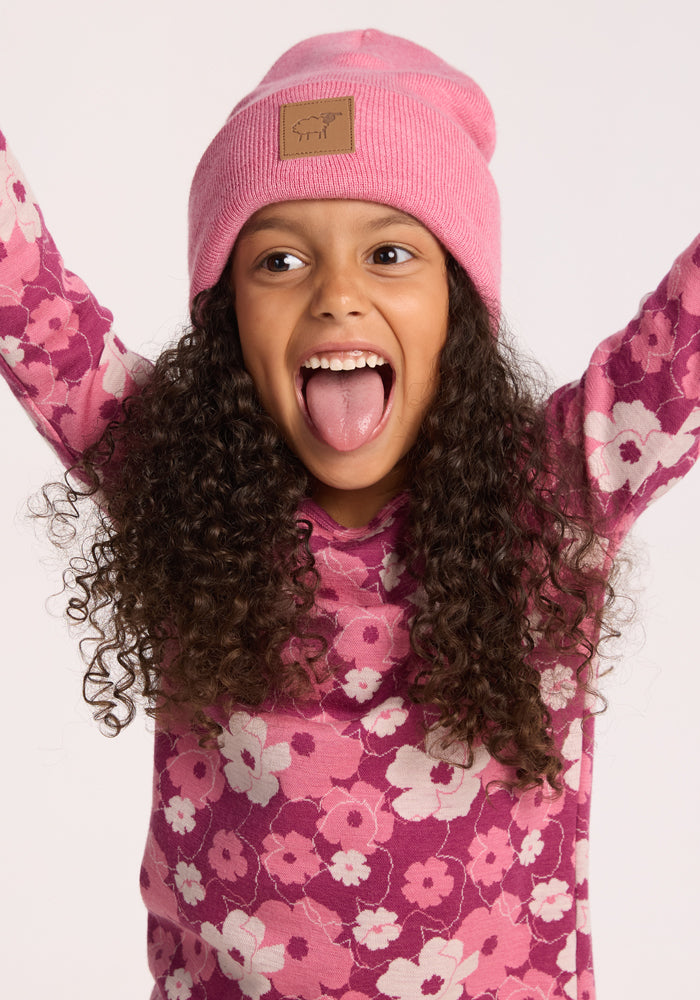 A smiling young girl with curly hair playfully sticks out her tongue, wearing the Woolx Kids Baylor Beanie - Pinky Promise and a pink floral shirt, with arms raised against a light background.
