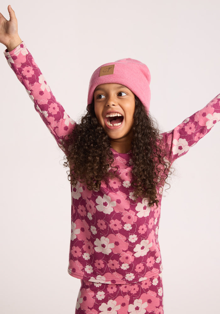 A young girl with long curly hair smiles widely, arms raised in excitement, wearing the Woolx Kids Baylor Beanie - Pinky Promise against a plain background.