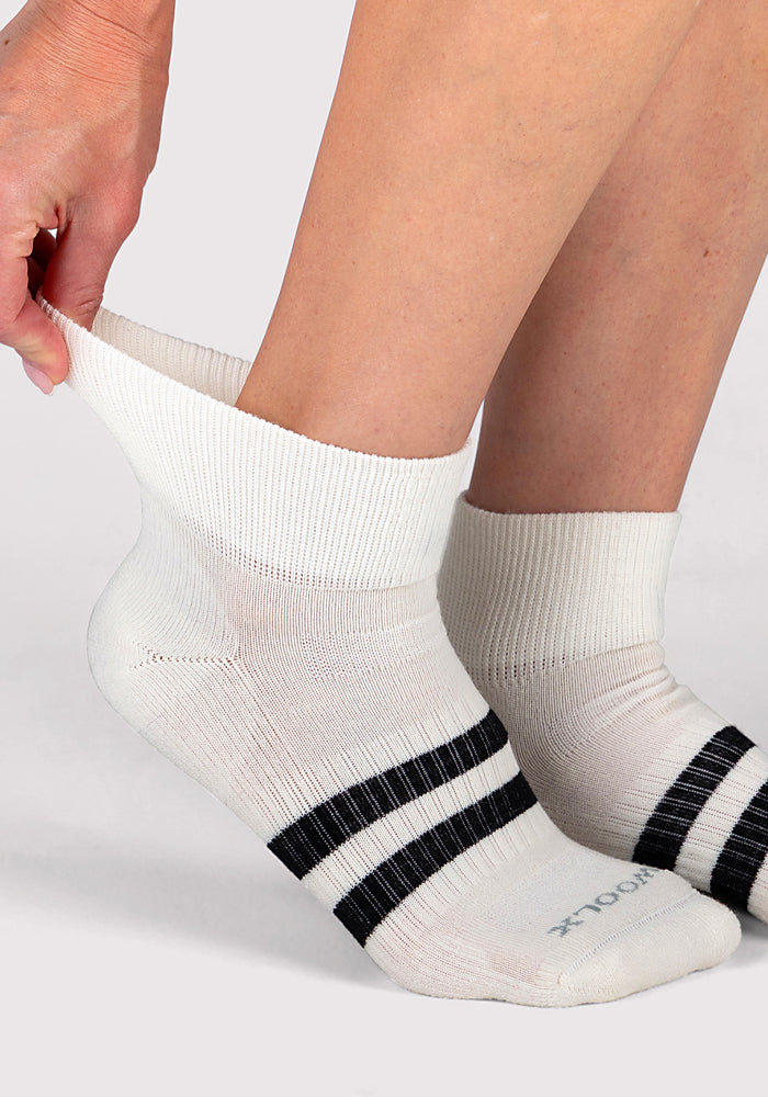 A person pulls up a Woolx Womens Rambler Ankle Sock in Polar White, featuring two black stripes, while their other foot already wears the matching sock against a plain light background.