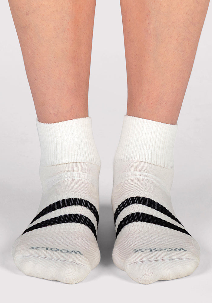 A person wears Woolx Womens Rambler Ankle Sock in Polar White, featuring black stripes and "WOMEN" on the toes, standing on a light gray surface for superior moisture management.