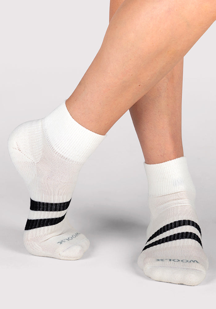 A person wearing Woolx Womens Rambler Ankle Sock in Polar White, featuring two black stripes near the toes and designed for moisture management, stands with one foot crossed over the other against a plain background.