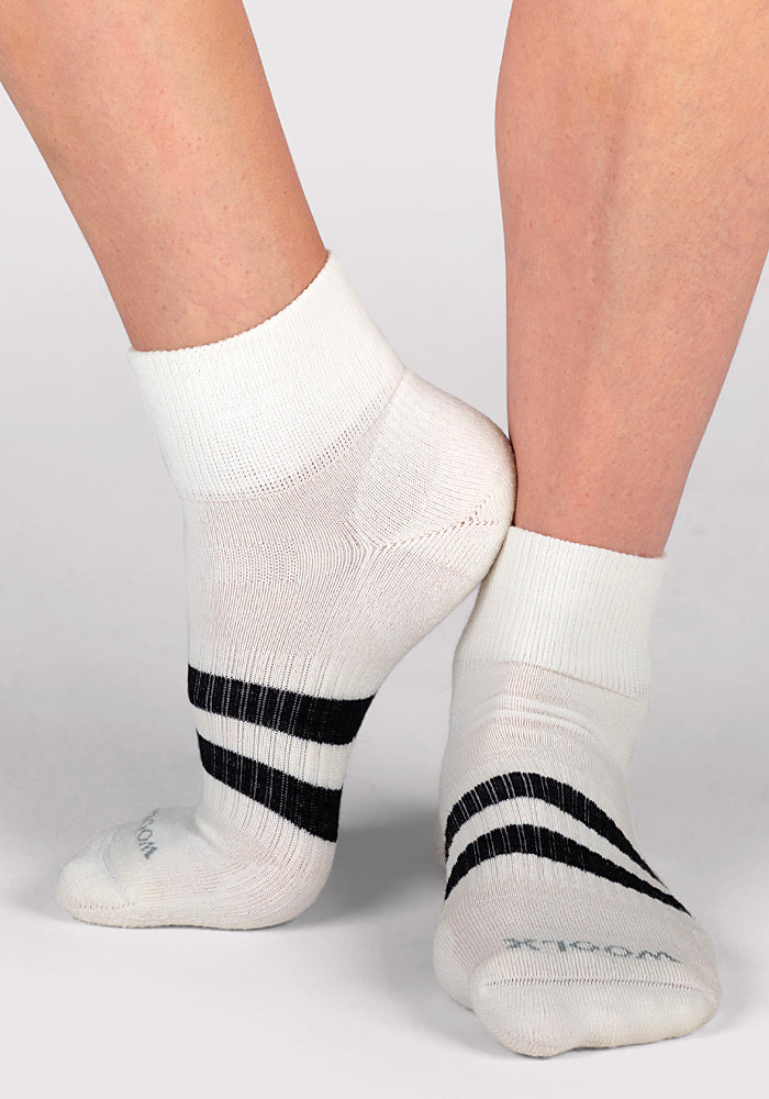 A close-up of two feet in Woolx Womens Rambler Ankle Socks - Polar White, featuring two black stripes near the toes, highlights their comfort and moisture-wicking Merino wool blend on a light surface with one heel slightly raised.