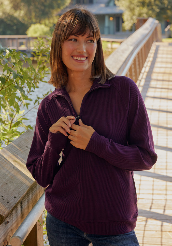 A woman with brown hair smiles on a sunlit wooden bridge, wearing the Woolx Quinn Ribbed 1/4 Zip in Majolica Blue. Trees and a house can be seen in the background.
