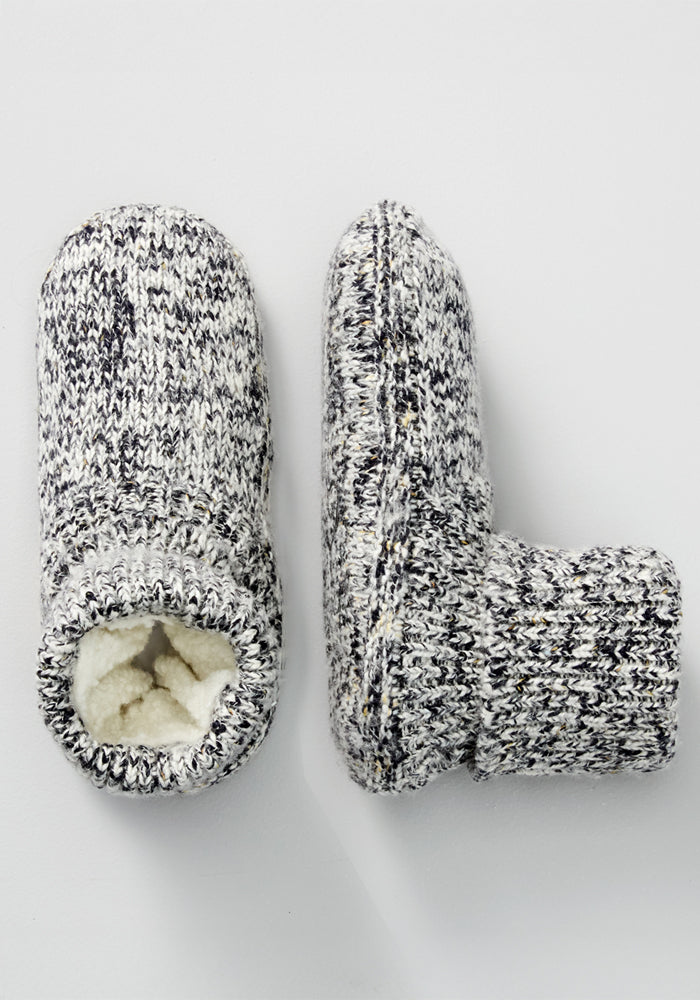 The Woolx Women's Gripper Slipper Booties - Speckled Stone feature cozy speckled knit in black and white, soft sherpa lining, and non-slip sole grippers. One bootie stands upright while the other rests on its side to show the sole.